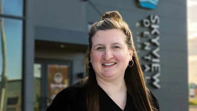 Smiling woman with long brown hair and hoop earrings standing outside Skyview Dental clinic in daylight.