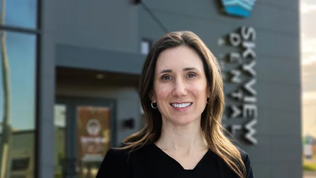 Smiling woman with brown hair stands outside Skyview Dental office building in natural light.