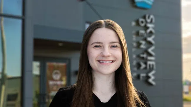 Smiling young woman with long brown hair standing outside a modern building with Skyview Dental sign.