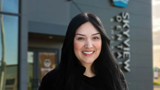 Smiling woman with long black hair stands outside Skyview Dental with building and logo in background.