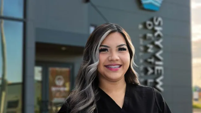 Smiling woman with long wavy hair and braces wearing black scrubs outside Skyview Dental Clinic.