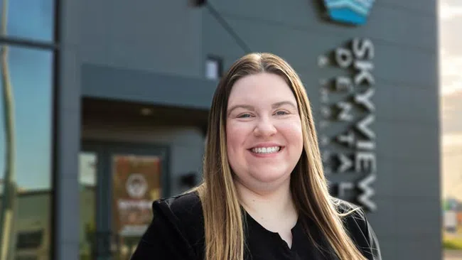 Smiling woman with long hair wearing black scrubs standing outside Skyview Dentistry building.