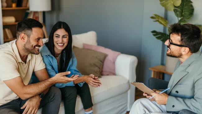 Couple consulting a smiling male therapist in a cozy living room with clipboard and conversation.