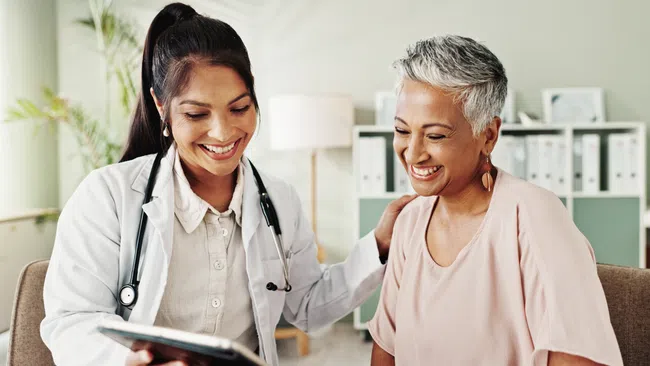 Female doctor showing a tablet and smiling with a happy middle-aged female patient in a medical office.