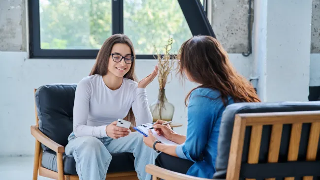 Young woman in casual clothes holding phone in therapy session with female counselor taking notes in bright office