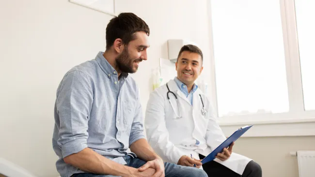 Male patient and doctor consulting in medical office with clipboard, smiling and discussing health.