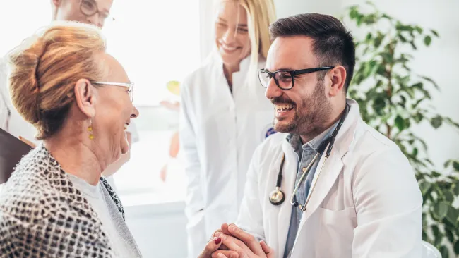 Friendly male doctor holding elderly patient's hands with smiling medical staff in the background.
