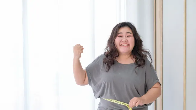 Happy plus-size woman measuring waist with tape measure, showing fitness progress and body positivity indoors.