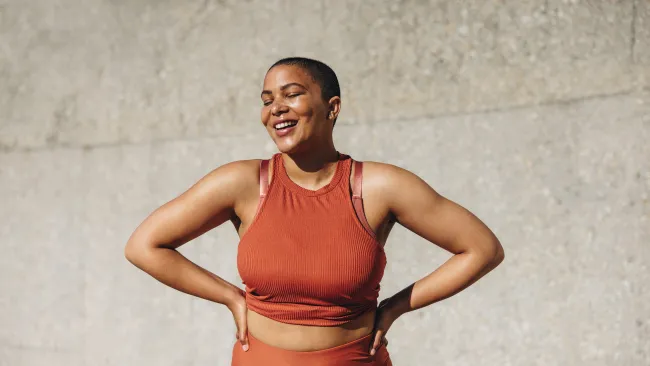 Smiling woman in orange athletic wear posing with hands on hips against concrete wall background