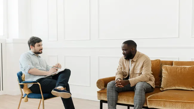 Two men engaged in a serious conversation in a bright room with modern furniture, one seated on a chair and the other on a sofa.