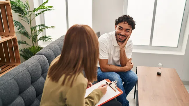 Young man smiling during a counseling session with a woman holding a clipboard on a gray sofa in a bright room