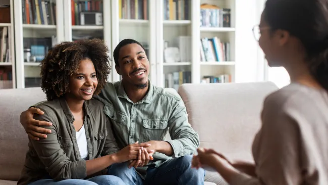 Happy couple sitting on a couch holding hands talking with a counselor in a home library setting.
