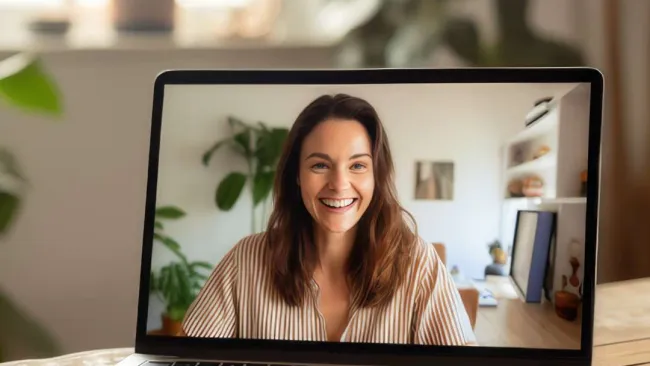 Smiling woman in striped shirt on video call displayed on laptop screen in home office setting.