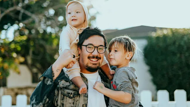 Father smiling and holding two young children outdoors near a white fence and trees in daylight.