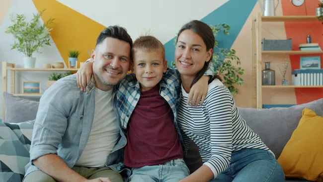 Smiling family of three sitting on a couch with colorful geometric wall and plants in the background