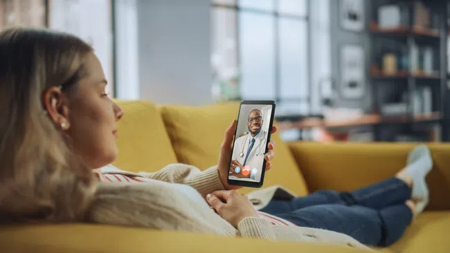 Woman lying on yellow couch having a video call with a doctor on her smartphone at home