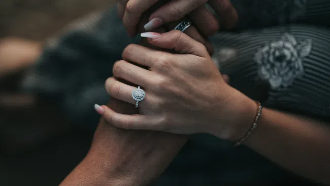Close-up of two people holding hands, featuring an engagement ring and floral patterned clothing.
