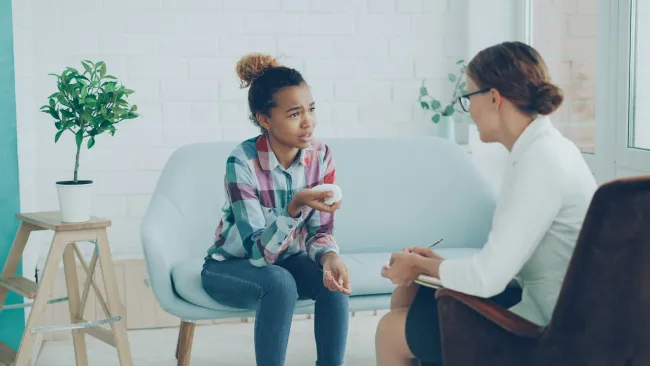 Teenage girl talking to a female therapist in a bright, modern counseling office with a potted plant.