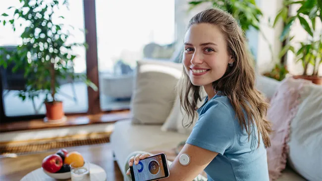 Smiling woman sitting on a couch using a glucose monitor app with a sensor on her arm in a bright living room.