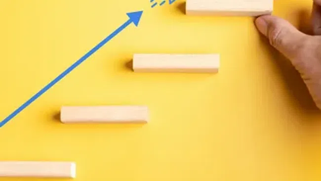 Hand placing wooden block on ascending steps with blue arrow, rocket icon, and red target symbol on yellow background