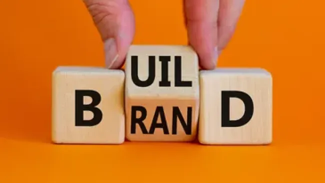 Hand arranging wooden blocks spelling build and brand on orange background representing growth and marketing.