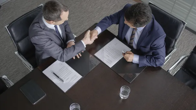 Two businessmen in suits shaking hands over signed contracts at a meeting table with glasses of water