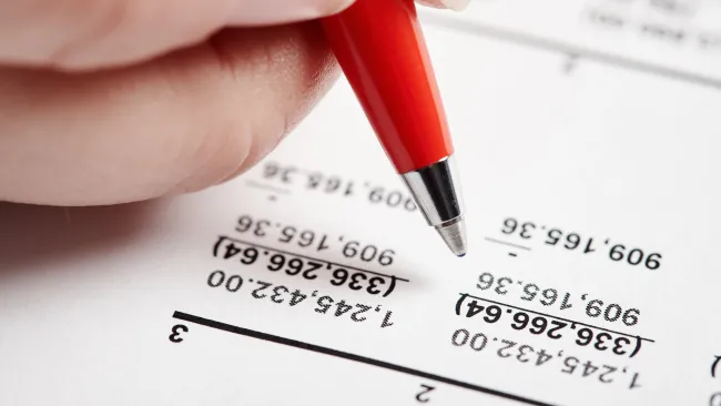 Hand holding red pen pointing to financial figures on printed document with highlighted numbers.
