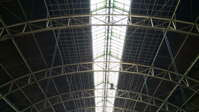 Interior view of a large industrial roof structure with metal beams and a central skylight.