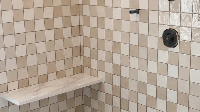 Beige tiled shower area with built-in corner bench and matte black fixtures including shower controls and spout.