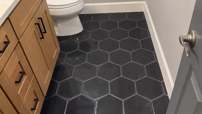 Modern bathroom with black hexagonal tile floor, wooden vanity, white toilet, and gray door.