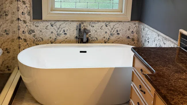 Modern bathroom with white freestanding tub, floral wallpaper, stained glass window, and wood cabinets.