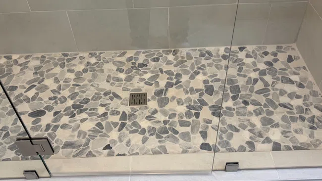 Modern shower floor with gray and white stone tiles and glass door featuring metal hinges and drain.