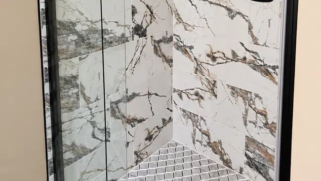 Modern shower with glass door, marble-patterned wall tiles, and mosaic floor tiles with floor drain.