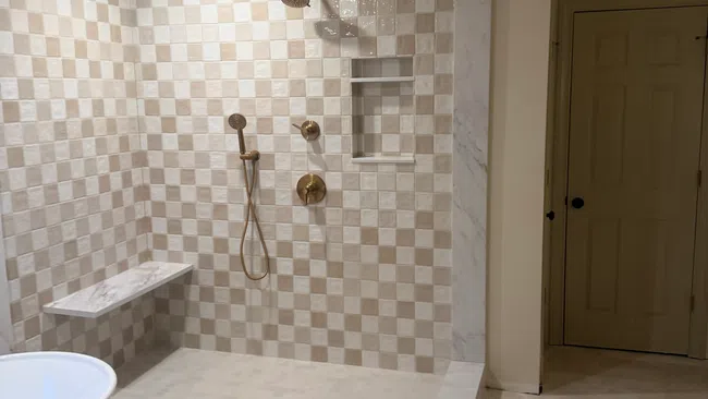 Modern walk-in shower with beige and white checkerboard tiles, bronze fixtures, built-in shelf, and marble bench.