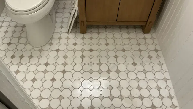 Bathroom floor with white circular tiles and tan grout next to a white toilet and wooden cabinet.