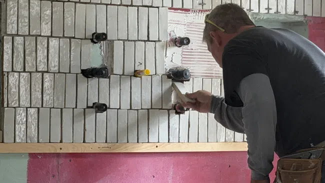 Worker installing rectangular white tiles on wall above a pink and green painted surface using spacers and adhesive.