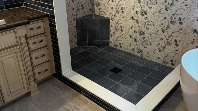 Modern bathroom shower with dark tiles, floral wallpaper, built-in niche, and beige vanity with granite countertop.