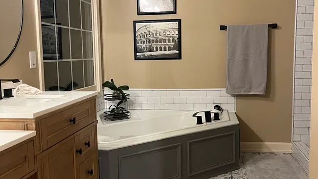 Modern bathroom with patterned tile floor, corner bathtub, wooden vanity, and travel-themed black and white wall art.