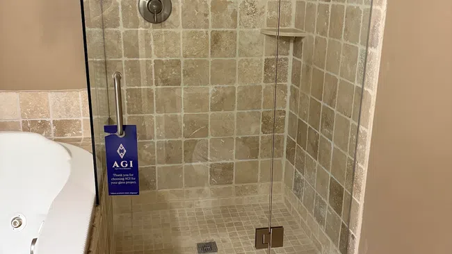 Glass shower enclosure with beige tile walls, built-in shelves, and a brown bath mat next to a white bathtub.