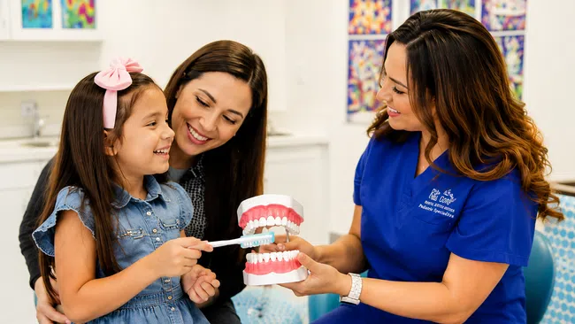 Dental hygienist teaching a young girl and her mother how to brush teeth using a model in a dental clinic.
