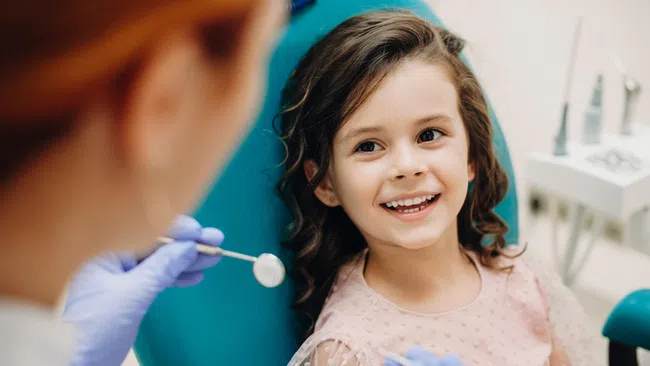 Smiling young girl sitting in dental chair during checkup with dentist holding dental mirror and explorer.