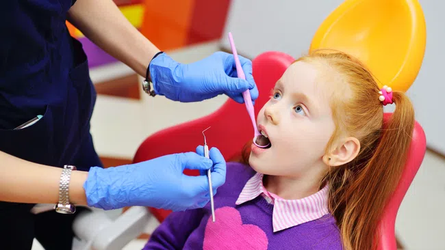 Child with red hair in a purple sweater having a dental checkup by a dentist wearing blue gloves.