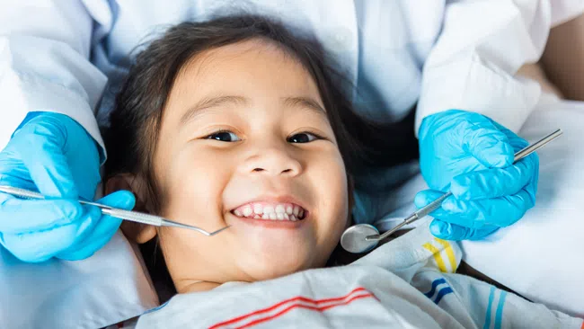 Smiling child receiving dental checkup with dentist wearing blue gloves and holding dental tools.