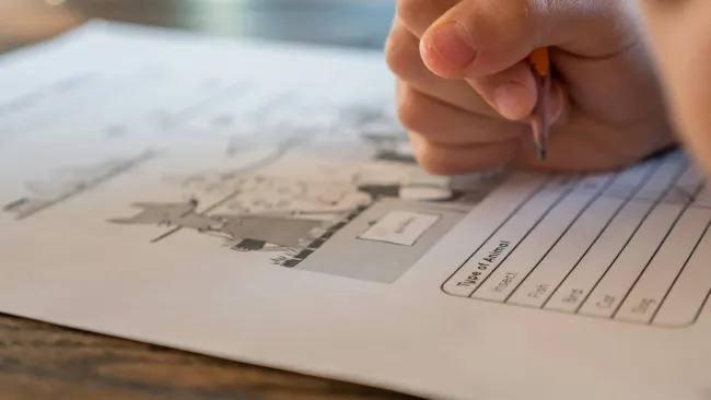 Child's hand holding a pencil writing on an animal-themed worksheet on a wooden table.