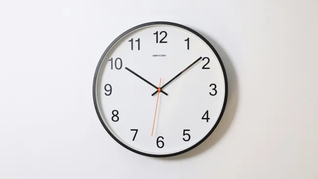 Round wall clock with black frame showing time 10:10 on a white background with orange second hand