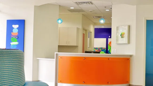 Bright modern reception area with orange desk, blue seating, colorful ceiling panels, and clean white walls.