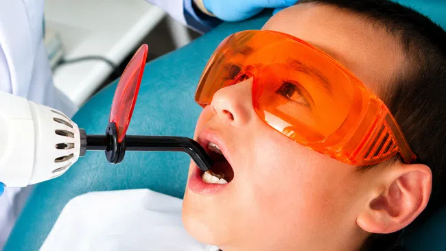 Child wearing orange protective glasses receiving dental laser treatment from dentist in clinic.