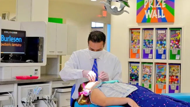 Dentist treating a young girl wearing pink headphones in a bright pediatric dental clinic with a colorful prize wall.