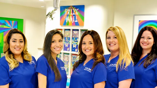 Five female dental professionals smiling together in blue scrubs inside a colorful pediatric dental office.