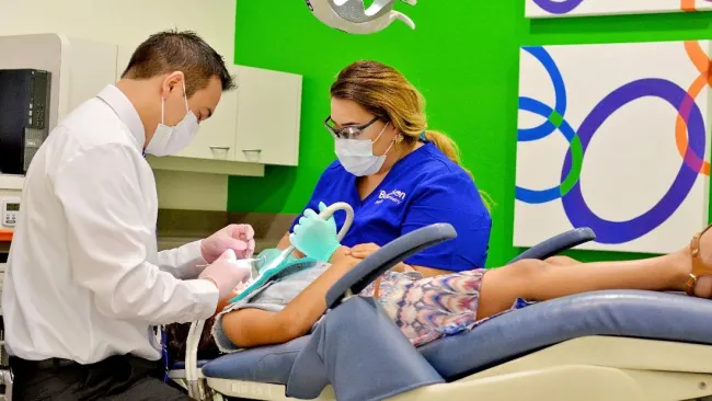 Dentist and assistant wearing masks treating a patient lying on a dental chair in a modern clinic.